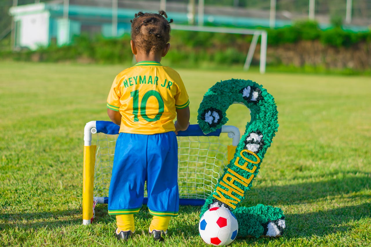 A young child wearing a Neymar Jr. jersey stands by a mini soccer goal on a grassy field.